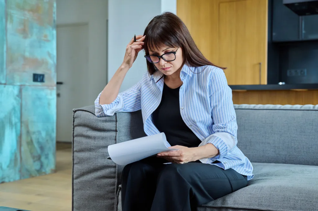 Woman sitting on a couch reviewing paperwork with a concerned expression.